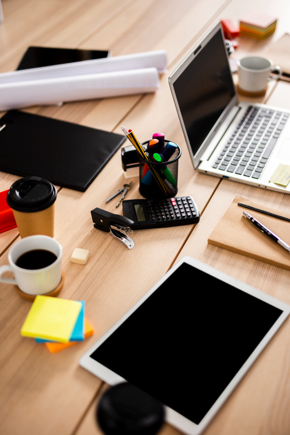 Computer portatile e tablet su una scrivania, tra calcolatrice, post-it, matite e tazza di caffè