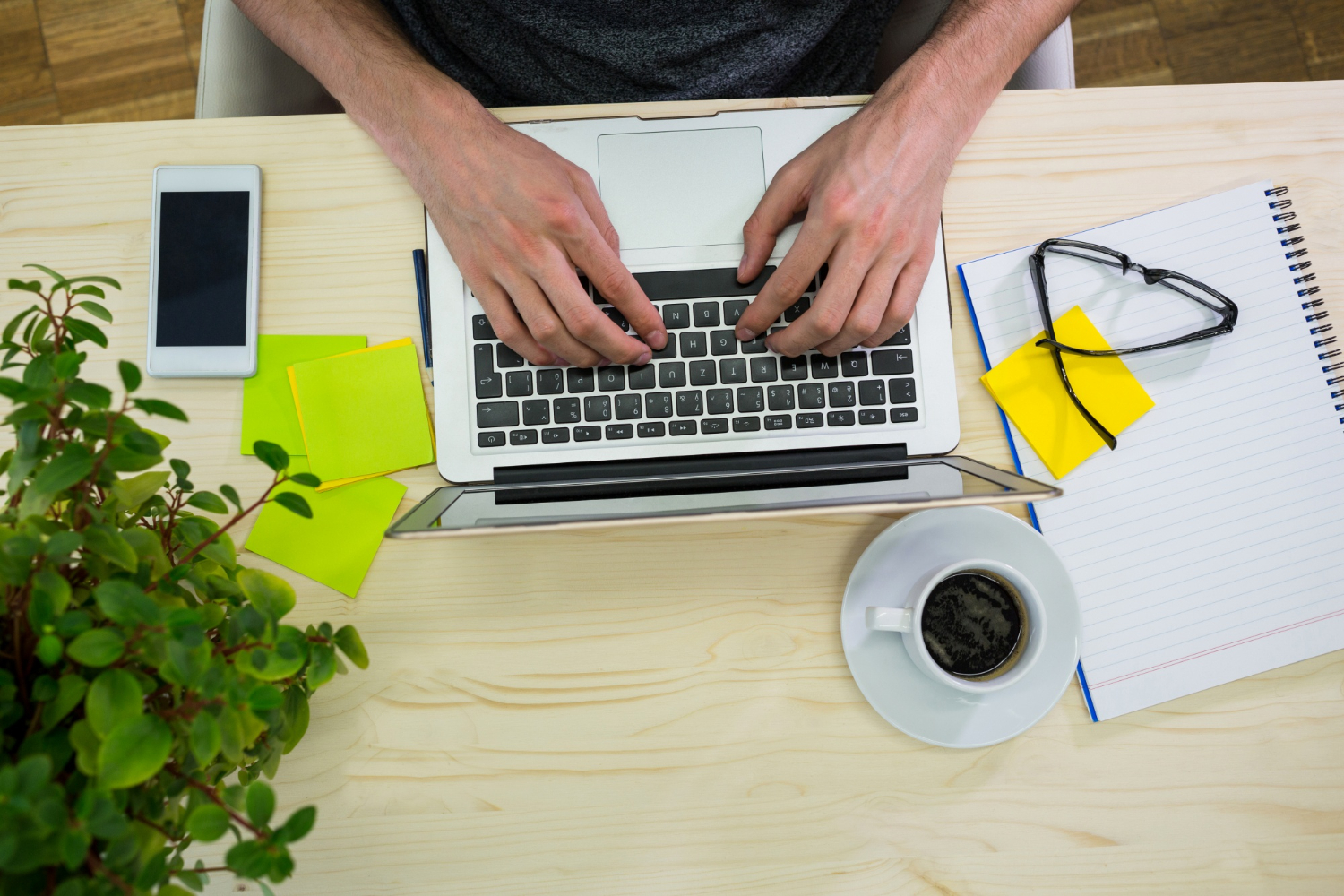 Mani di uomo sulla tastiera di un portatile, con a fianco post-it, occhiali da vista e tazza di caffè