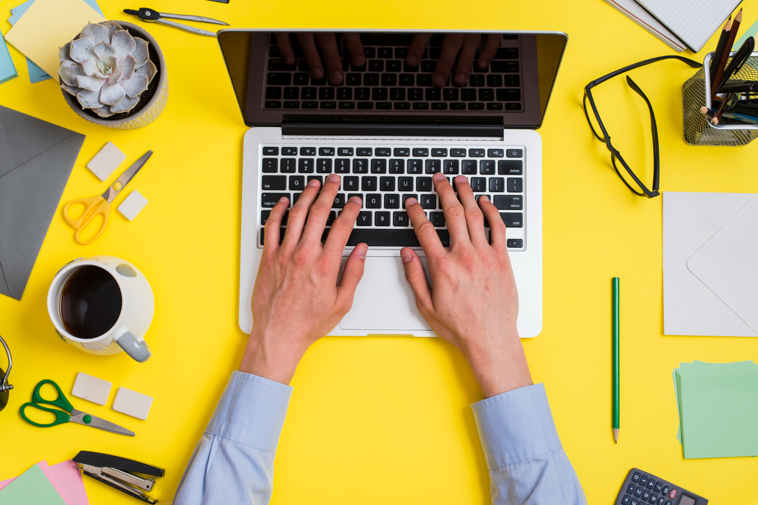 Mani di uomo su un laptop, su una scrivani a con penne, forbici, post-it, occhiali da vista e tazze
